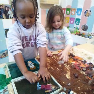 Two children engaging in sensory play with trays of water and mud, exploring textures with small figurines and natural materials.