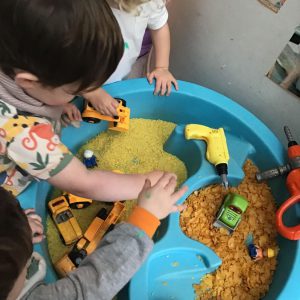 Children engaging in sensory play with a blue activity table filled with colored rice, toy construction vehicles, plastic tools, and cereal pieces.