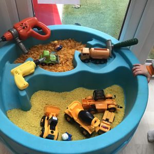 Blue sensory activity table filled with toy construction vehicles, tools, yellow rice, and cereal pieces, set up near a window with a 'Dream' construction-themed sign.