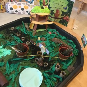 Blue sensory activity table filled with toy construction vehicles, tools, yellow rice, and cereal pieces, set up near a window with a 'Dream' construction-themed sign.