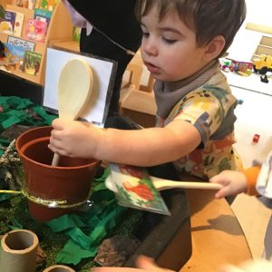 Child engaged in a sensory activity with a plant pot, wooden spoon, and greenery in a classroom setting