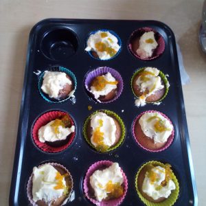 A tray of decorated cupcakes in colorful silicone liners, topped with cream and fruit jam.