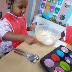 A child in a red apron scooping batter into colorful cupcake liners during a baking activity.