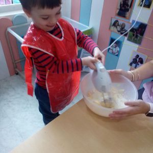 A smiling child in a red apron mixing batter with an electric hand mixer during a baking activity.