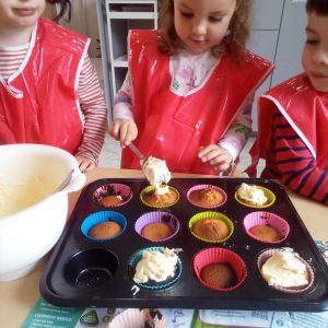 Children in red aprons decorating cupcakes with cream during a baking activity.